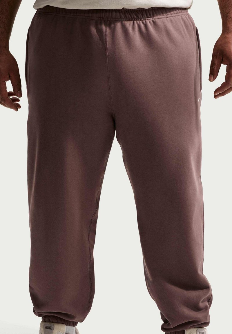 Persoon draagt losse bruine joggingbroek en een wit shirt, staat met ontspannen armen; onderaan zijn Nike-sneakers zichtbaar.