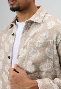 Bruin patroon overhemdjack gemaakt van zachte stof, voorzien van een knoopsluiting, borstzak en paisley ontwerp in wit.