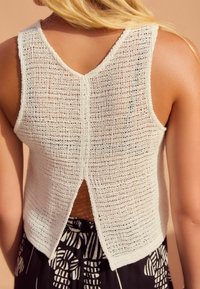 Hvid strikket tanktop med V-udskæring bagpå, der har en kløft i den nederste del af ryggen. Teksturen er åbenstrikket, hvilket viser et lagdelt look med mønstre nedenfor.