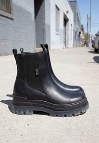 Paire de bottines en cuir noir avec des côtés élastiques côtelés et des semelles épaisses en caoutchouc, posée sur un trottoir urbain ensoleillé.