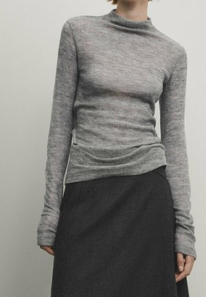 Vrouw die een aansluitende grijze coltrui met lange mouwen en een donkergrijze hooggetailleerde rok draagt tegen een effen achtergrond.