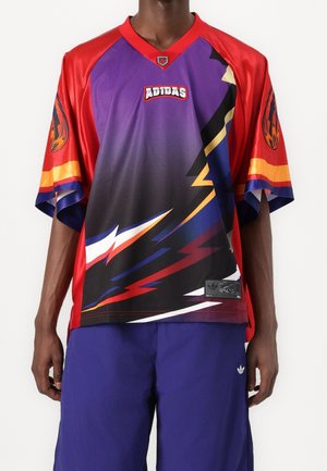 Osoba nosząca kolorową sportową koszulkę marki Adidas z czerwonymi rękawami oraz wzorem piorunów w kolorach fioletowym, czarnym i żółtym, zestawioną z fioletowymi szortami.