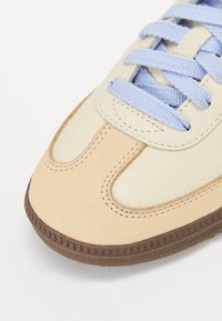 Närbild av en beige och krämfärgad sneakersko med ljusblå snören och en texturerad brun gummisula.