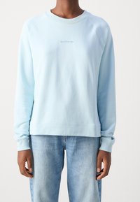 Sudadera azul clara con cuello redondo, mangas raglán y puños acanalados. Logotipo de la marca centrado impreso en texto sutil. Tejido suave y texturizado.