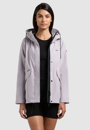 Naine, kes kannab heledat halli kapuutsiga jopet musta pluusi ja mustade lühikeste pükstega, seisab ühevärvilise tausta ees.