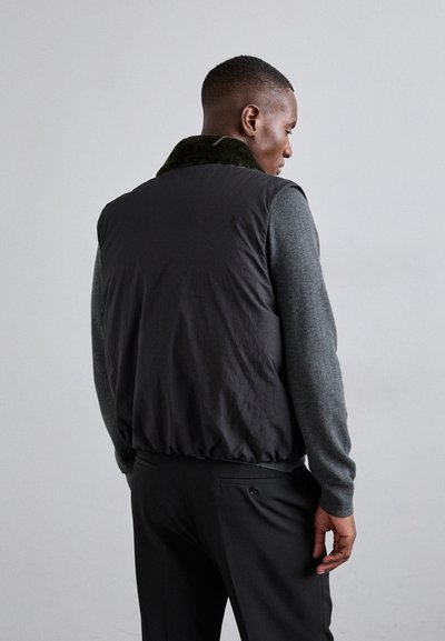 Črna prešita jakna z zelenim krznom na ovratniku, nosi se čez sivo pulover. Ima elastičen rob in gladko površinsko teksturo.