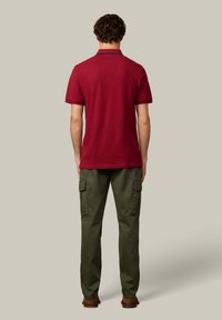 Czerwona koszulka polo z krótkim rękawem, widziana od tyłu, zestawiona z zielonymi spodniami typu cargo. Strój ma prosty design, bez wzorów.