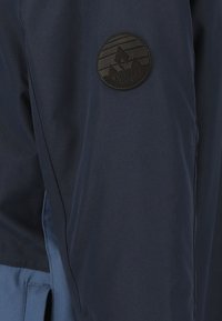 Mörkblå jacka med en texturerad tyg, med en svart cirkulär logotypapplikation som visar berggrafik på överarmen.