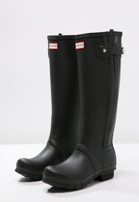 Botas de lluvia de goma negras con un diseño alto, que presentan un detalle de hebilla, suela texturizada y logotipos en la parte superior. Acabado suave y mate.
