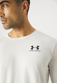 Bijela košulja dugih rukava od pamuka, sa crnim logotipom Under Armour na gornjem lijevom prsnom dijelu, okruglim izrezom i glatkom teksturom.