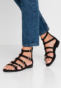 Musta mokkasandaalit, joissa on useita hihnoja ja hopeansävyisiä solkia. Pohja on tasainen, mikä tekee siitä rentoa muotoilua sopivaa lämpimälle säälle.