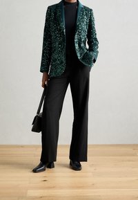 Smaragdgrön paljettblazer med satinkrage, matchad med svarta vida byxor och svarta klackiga ankelstövlar, håller en liten svart v äska.