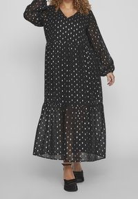 Vestido maxi negro con mangas largas de tul, escote en V y patrones en forma de lágrima blancos. Presenta una falda fluida con un dobladillo en capas. Zapatos negros.