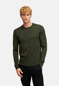 Grüner Strickpullover mit langen Ärmeln, rundem Ausschnitt, geripptem Saum und strukturiertem Oberflächen, kombiniert mit schwarzen Hosen.
