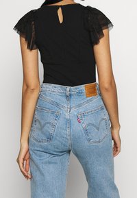 Czarny kombinezon z przezroczystymi koronkowymi, falbaniastymi rękawami i wycięciem na plecach, zestawiony z jasnoniebieskimi dżinsami z wysokim stanem, wyposażonymi w skórzaną naszywkę i zakrzywione kieszenie.