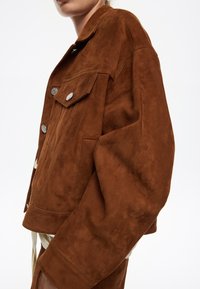 Giacca in suede marrone con vestibilità rilassata, tasche frontali e bottoni argentati. Texture liscia e maniche corte, leggermente accorciate.