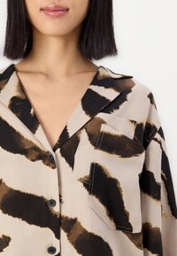 Camicia beige chiaro con motivo astratto marrone scuro e nero, dotata di colletto, chiusura a bottoni e taschino sul lato sinistro del petto.