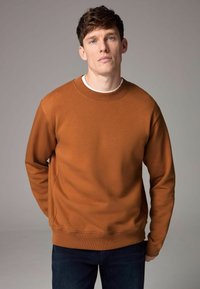 Sudadera de bronce con cuello redondo y puños de canalé, hecha de una tela suave; combinada con jeans azul oscuro sobre un fondo neutral.