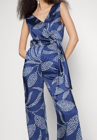 Femme portant une combinaison sans manches en satin bleu marine avec motif feuilles blanches à pois et détail noué sur le côté, debout les mains sur les hanches.