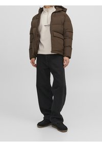 Chaqueta de plumas marrón con capucha, cierre de cremallera y textura acolchada. Usada sobre una sudadera ligera y combinada con jeans de pierna ancha negros y botas negras.