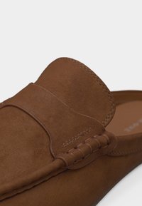 Mocassin en cuir suédé marron à enfiler avec des détails cousus et un bout arrondi, montré en gros plan sur un fond uni.