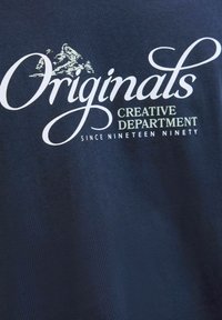 Tumesinine puuvillane t-särk, millel on valgega kirjutatud tekst "Originals" ja "Creative Department" koos mägiga, sile tekstuur ja avar lõige.