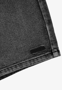 Czarna tkanina dżinsowa o gładkiej strukturze, z czarną metką blisko dolnej krawędzi. Tkanina ma delikatny, zużyty wygląd z widocznymi szwami.