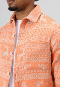Oranje overhemd met patroon en een zak, gemaakt van textuurstof met hertenmotieven en geometrische ontwerpen, met knopen aan de voorkant en een kraag.