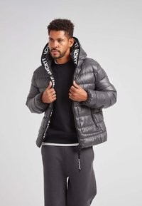 Chaqueta acolchada gris con cierre frontal, puños elásticos y capucha forrada con tela de marca. Textura suave, corte cuadrado y diseño acolchado.