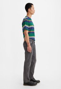 Camiseta a rayas en azul, verde y naranja con mangas cortas, combinada con jeans grises. El modelo lleva zapatos negros y tiene detalles en las manos con patrones.