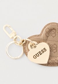 Přívěsek na klíče se zlatým srdíčkem s vyraženým nápisem "GUESS", připevněný k texturovanému béžovému koženému ozdobnému srdíčku a kovov�ému zacvakávacímu klipu.