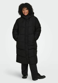 Melnā pūkaina jaka ar kapuci, ar garu, kasti līdzīgu dizainu, lielām tādām kā šūtiem laukumiem un sānu kabatām. Valkāta pār tumšām biksēm.