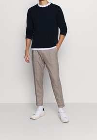 Marineblauer Pullover, darunter ein weißes Rundhals-Shirt, kombiniert mit hellbraunen, karierten, schmal zulaufenden Hosen und weißen Sneakers, die einen lässigen Look zeigen.