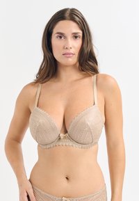 Reggiseno beige in pizzo con scollatura profonda, tessuto testurizzato, bordi ondulati e un piccolo fiocchetto al centro. Spalline regolabili incluse.