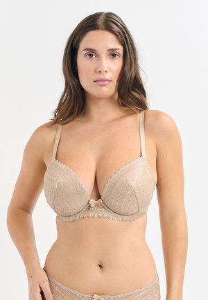 Reggiseno beige in pizzo con scollatura profonda, tessuto testurizzato, bordi ondulati e un piccolo fiocchetto al centro. Spalline regolabili incluse.