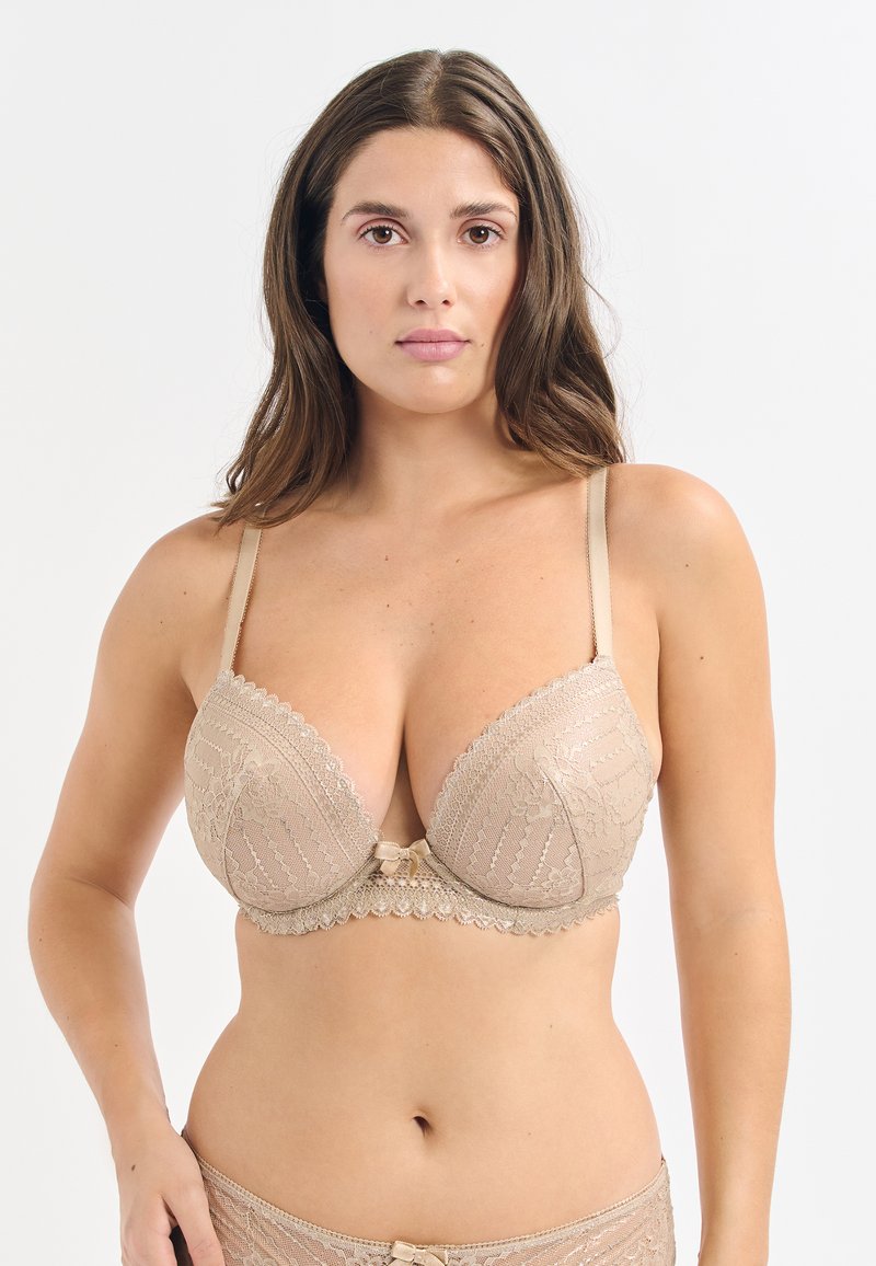 Reggiseno beige in pizzo con scollatura profonda, tessuto testurizzato, bordi ondulati e un piccolo fiocchetto al centro. Spalline regolabili incluse.