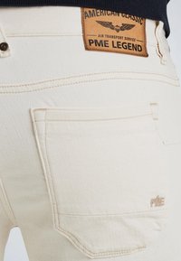 Jean en denim couleur crème avec une étiquette en cuir portant la mention "PME LEGEND". Les caractéristiques incluent des poches cousues et un tissu texturé.