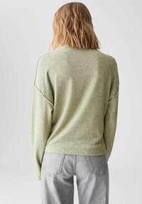 Blonde vrouw met haar rug naar de camera, gekleed in een lichtgroene lange mouwen trui en lichtgrijze spijkerbroek, tegen een effen lichte achtergrond.