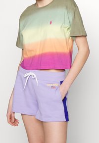 Person som bär en kortärmad, flerfärgad gradiente tshirt och ljuslila shorts med dragsko och "Polo Sport Ralph Lauren"-logotyp på fickan.