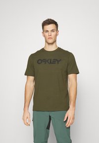 Olivenfarvet kortærmet T-shirt lavet af bomuld. Har et iøjnefaldende sort "OAKLEY" logo på tværs af brystet. Glat tekstur, klassisk pasform.
