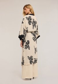 Dlouhý kimono v světle béžové látce s černým květinovým potiskem, má široké rukávy a kontrastní černou lemovku podél okrajů.