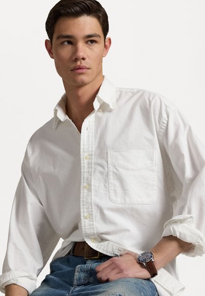 Camisa blanca de botones con un bolsillo, corte relajado y mangas arremangadas, combinada con unos jeans azul claro y un reloj de cuero marrón.