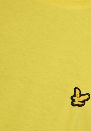 Tissu jaune avec un petit emblème d'oiseau jaune brodé, contouré de noir, près du coin inférieur droit.