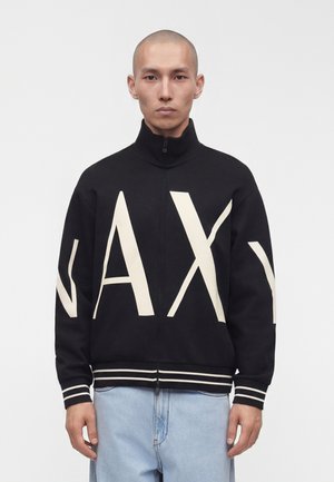 Hombre con cabeza rapada que lleva una chaqueta negra con cremallera con grandes letras blancas "A|X" y vaqueros azul claro, de pie frente a un fondo sencillo.