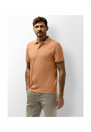 Man with short brown hair and beard wearing a light brown polo shirt and beige pants, standing with one hand in pocket against white wall.