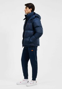 Granatowa kurtka puchowa z wysokim kołnierzem i małym logo, zestawiona z pasującymi spodniami dresowymi i białymi trampkami. Gładka tekstura, lekka konstrukcja.