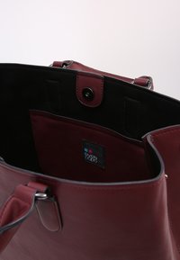 Bordeaux leren schoudertas met een zwarte interieur, drukknoopsluiting en een binnenzak met een label. Glad oppervlak en stevige handvatten.