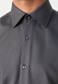 Homme portant une chemise de couleur gris foncé avec un col pointu et trois boutons noirs visibles attachés à l'avant.