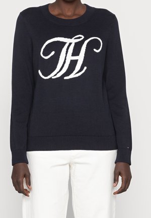 Ciemnogranatowy sweter z białym stylizowanym logo "TH" na piersi, noszony z białymi spodniami, ramiona swobodnie opuszczone wzdłuż ciała.