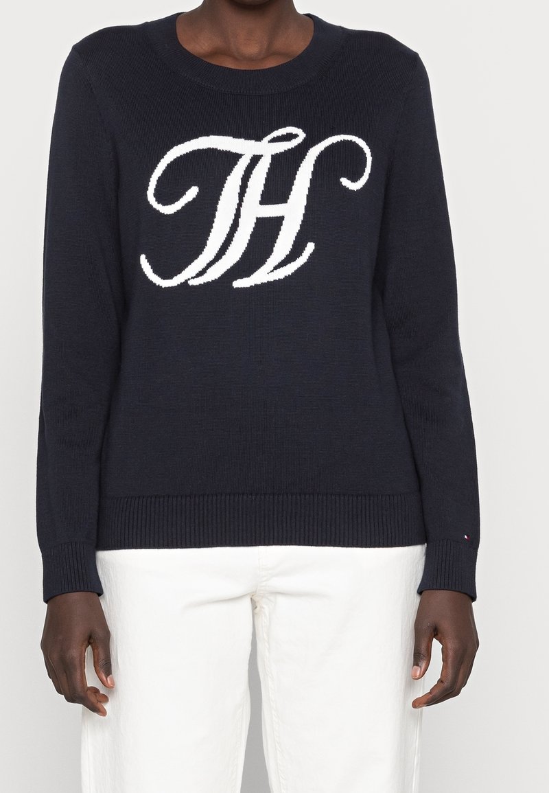 Pull marine foncé avec logo stylisé "TH" blanc sur la poitrine, porté avec un pantalon blanc, bras détendus le long du corps.